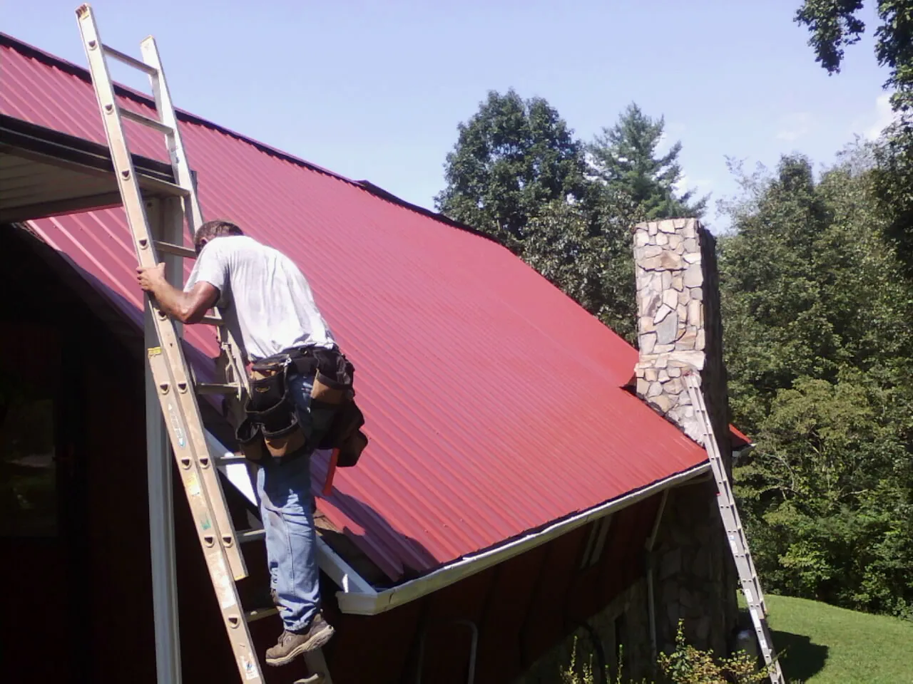 Roofer on a ladder beside a red metal roof and stone chimney — Soffit & Fascia Repair by Roofing Brothers in Nunn